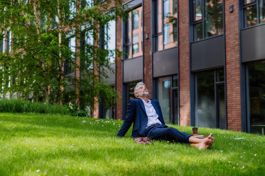 Ein Geschäftsmann, der barfuß auf einer neben dem Bürogebäude Wiese ruht, Work-Life-Balance-Konzept.