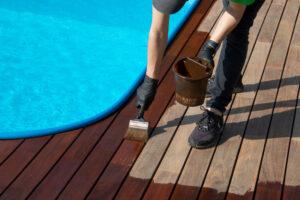 Arbeiter, der mit einem Pinsel braunes Holzschutzöl auf die Terrassendielen neben dem Pool aufträgt.