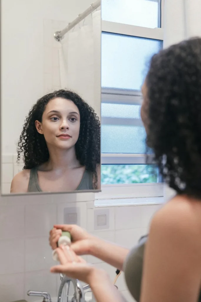 Junge Frau mit gelocktem Haar steht vor einem Badezimmerspiegel und bereitet sich darauf vor, Hautpflegeprodukte auf ihr Gesicht aufzutragen
