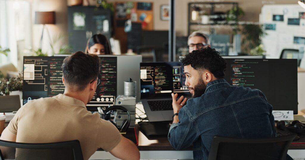 Zwei Maenner diskutieren Programmcode vor Monitoren in einem modernen Entwicklerteam im Buero