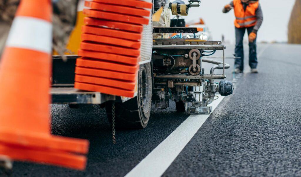 Detailaufnahme einer Maschine bei der Straßenmarkierung – weisse Linie wird aufgebracht, Arbeiter im Hintergrund