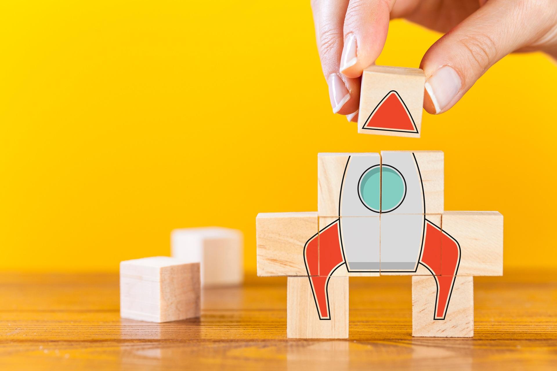 Hand baut mit Holzklötzen eine Rakete als Symbol für Start-up-Wachstum | Compliance Anwalt